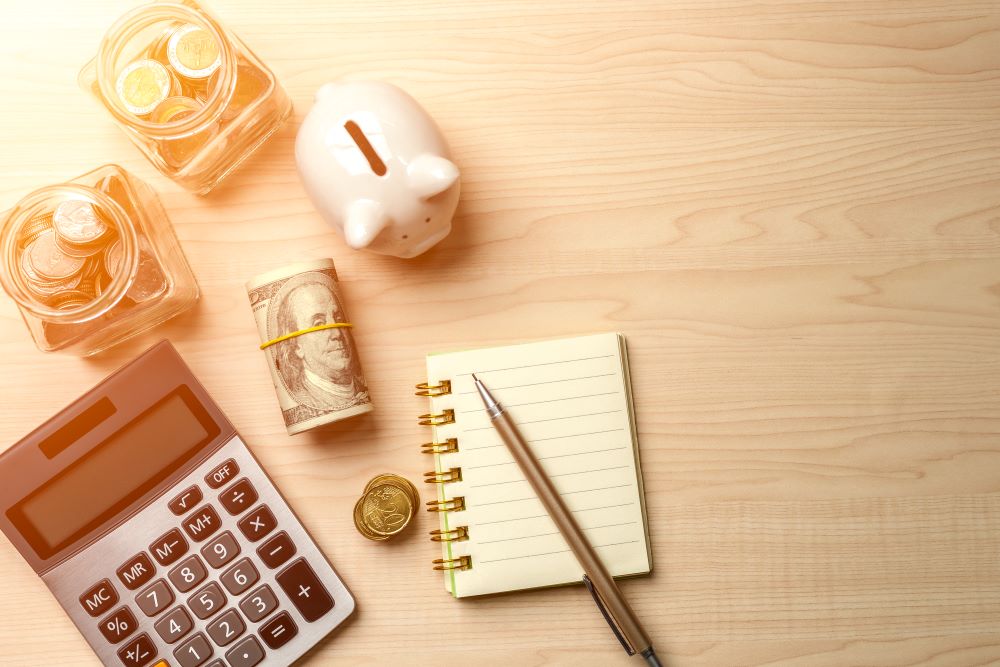 A calculator, notebook, piggy bank, and coin jars rest on a desk, indicating that with deliberation, you can potentially make your Self-Directed IRA a benefactor in your retirement planning.